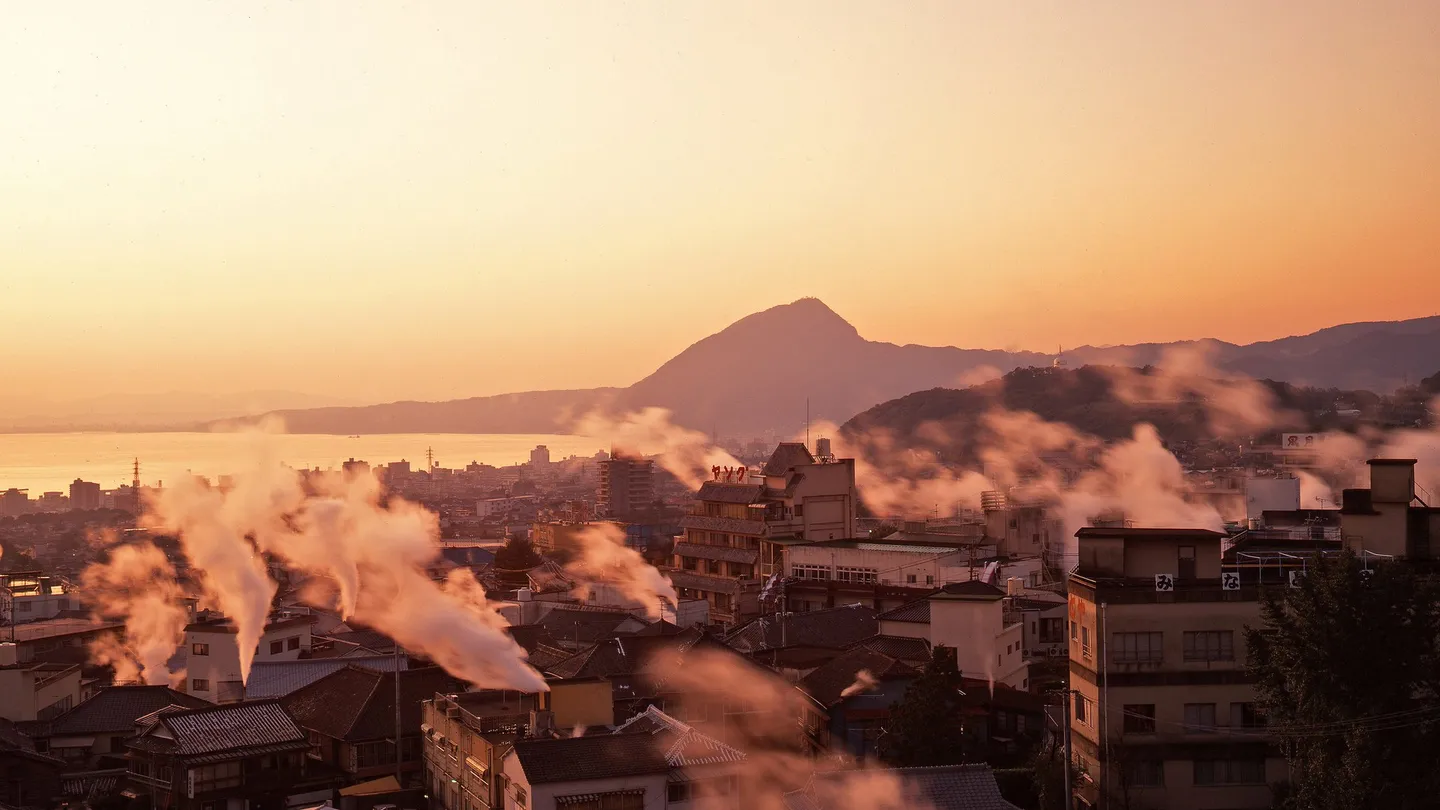 Vapor subindo das fontes termais entre as casas de Beppu, em Ōita, ao entardecer, com a baía ao fundo