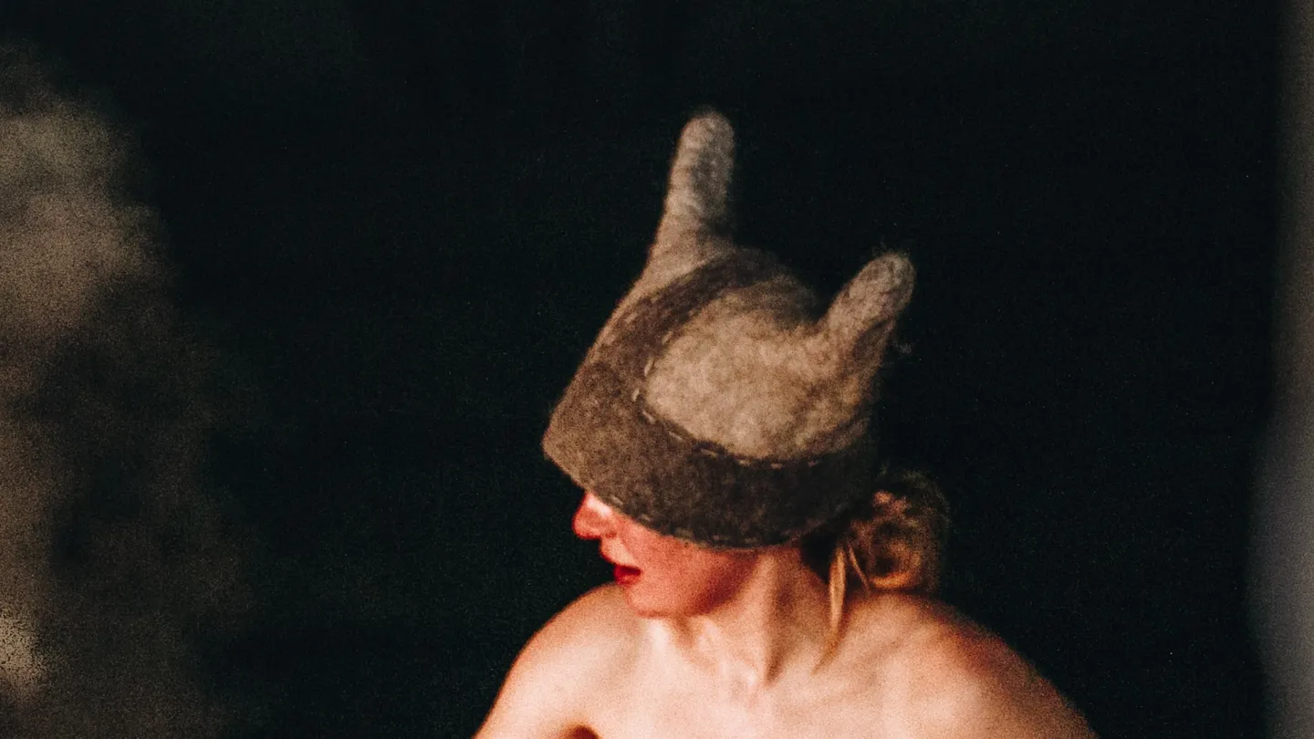 Banhista com um gorro de sauna de feltro com orelhas numa sauna de fumaça na Estônia, com o vapor subindo das pedras ao lado