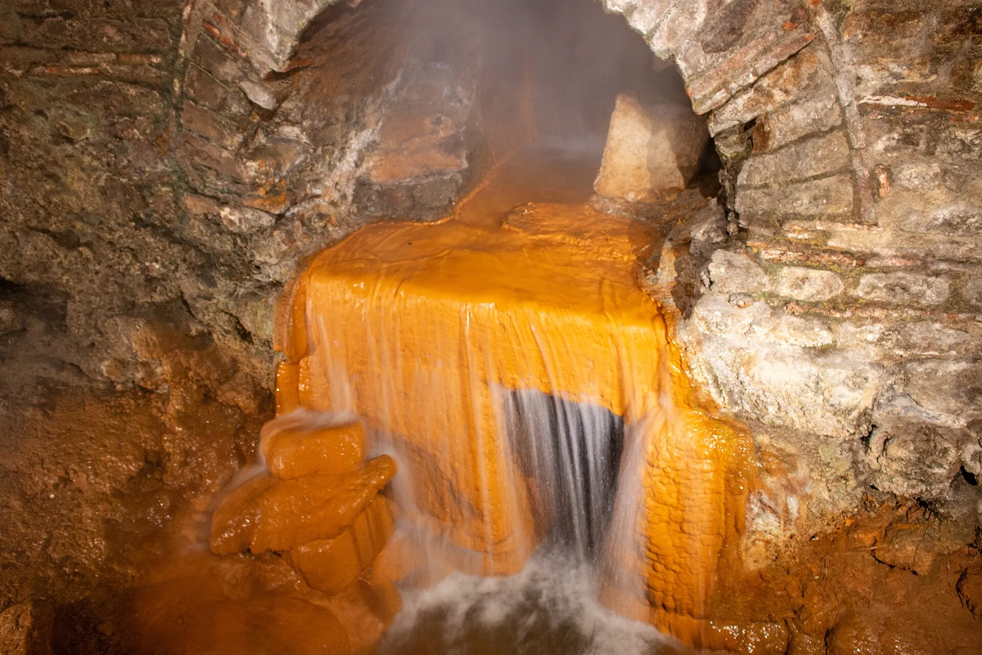 Água quente da Fonte Sagrada de Bath escorrendo por pedras tingidas de laranja pelo ferro, sob um arco de pedra das termas romanas