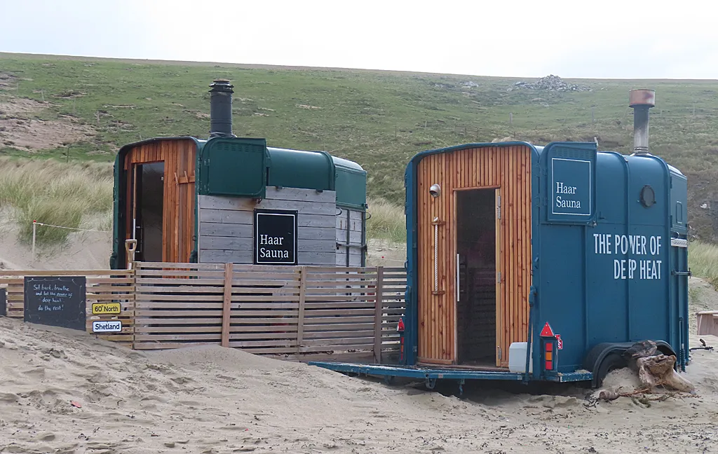 Duas saunas da Haar Sauna montadas em reboques de cavalo, uma verde e uma azul, sobre a areia de uma praia em Shetland