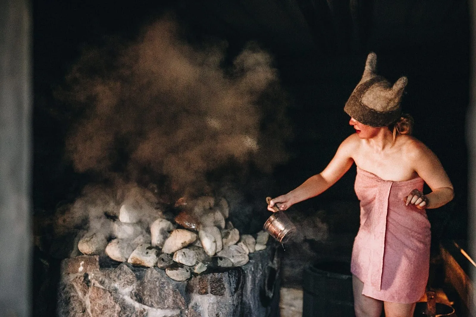 Mulher de toalha e gorro de feltro joga água sobre as pedras de uma sauna de fumaça na Estônia, com o vapor subindo