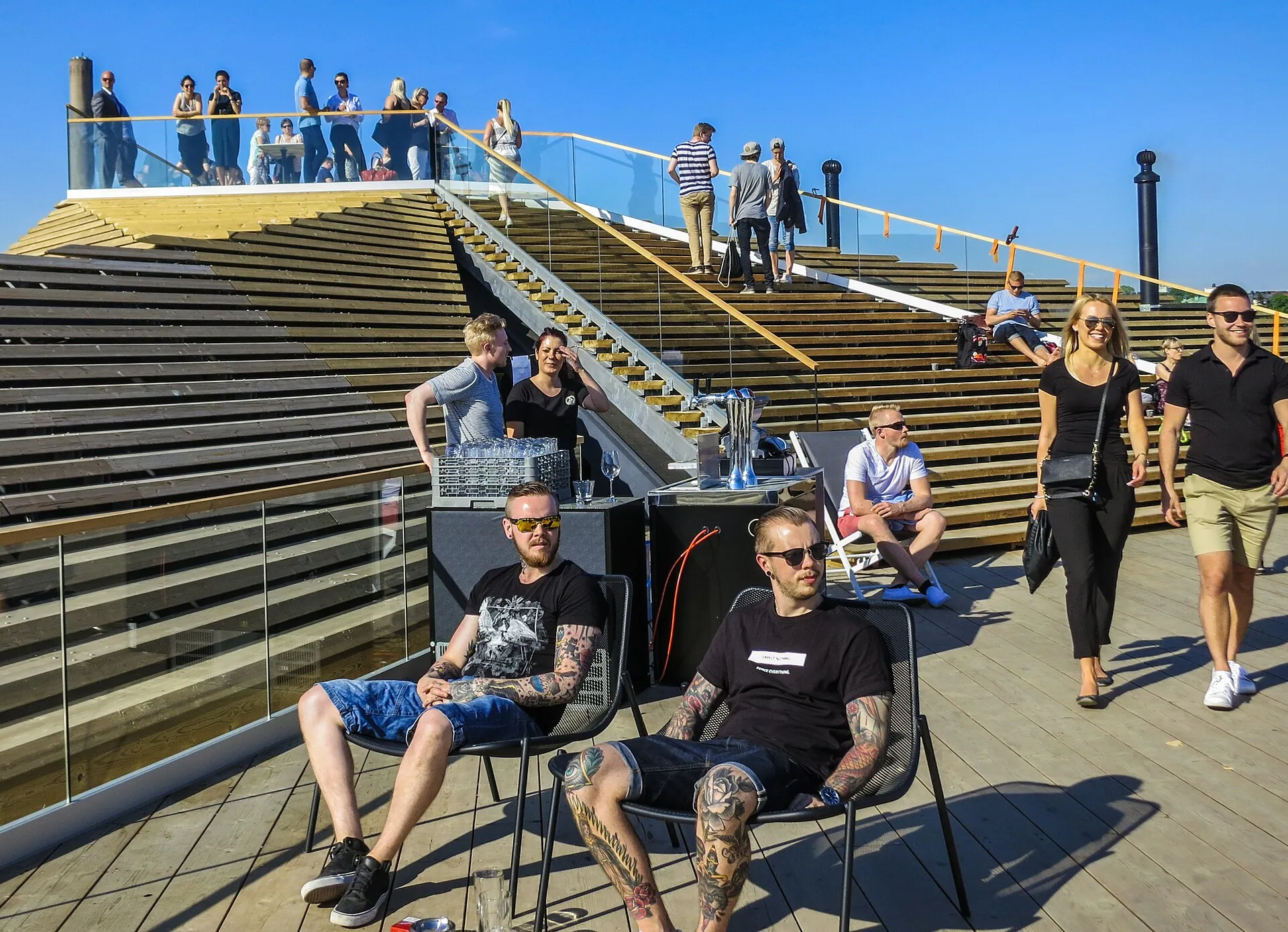 Terraço de madeira do Löyly, em Helsinque, com pessoas sentadas em cadeiras e em pé na escada do telhado escalonado, sob céu azul