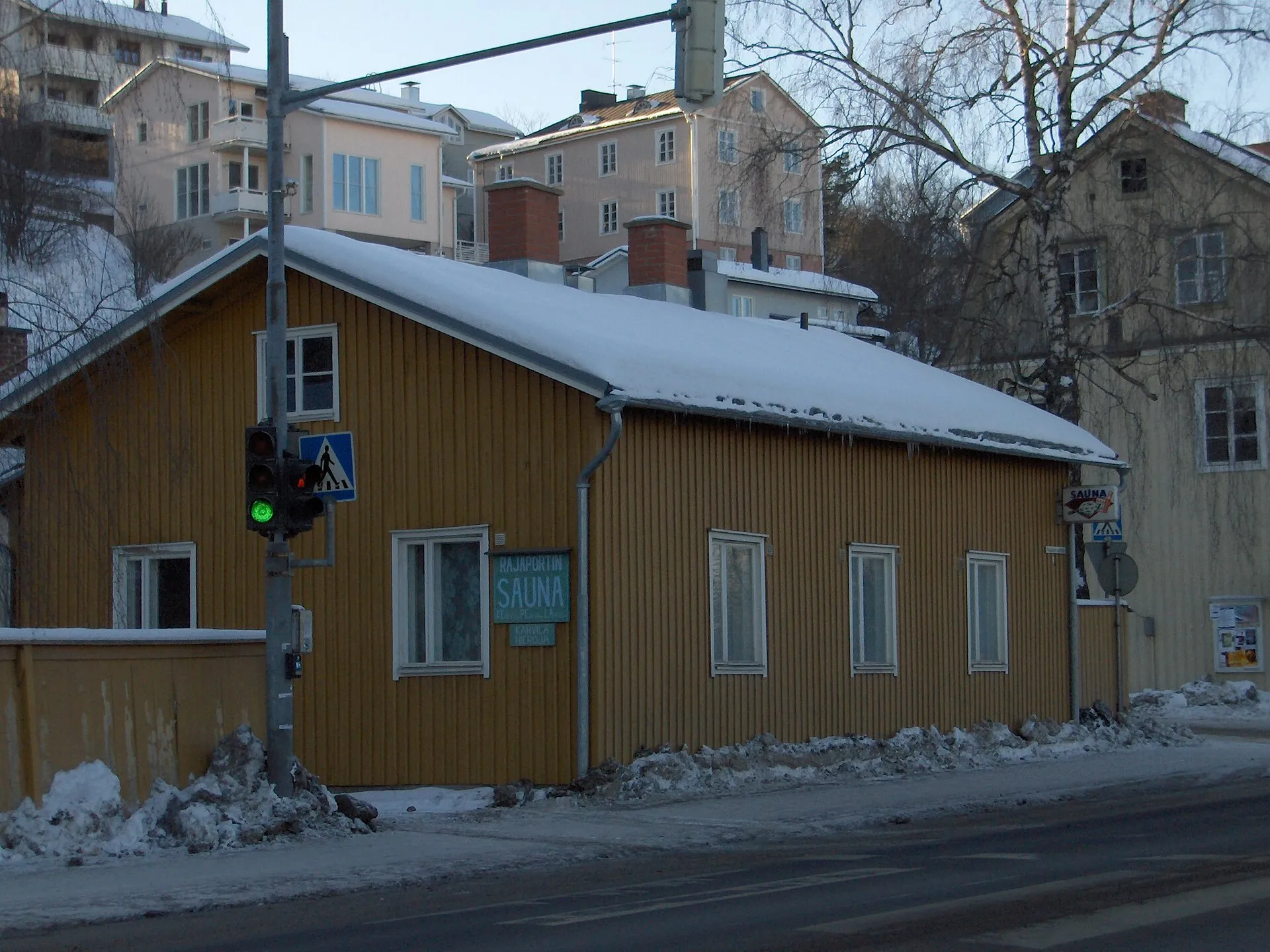 Prédio baixo de madeira amarela na esquina de uma rua nevada de Pispala, em Tampere, com a placa da sauna Rajaportti na fachada