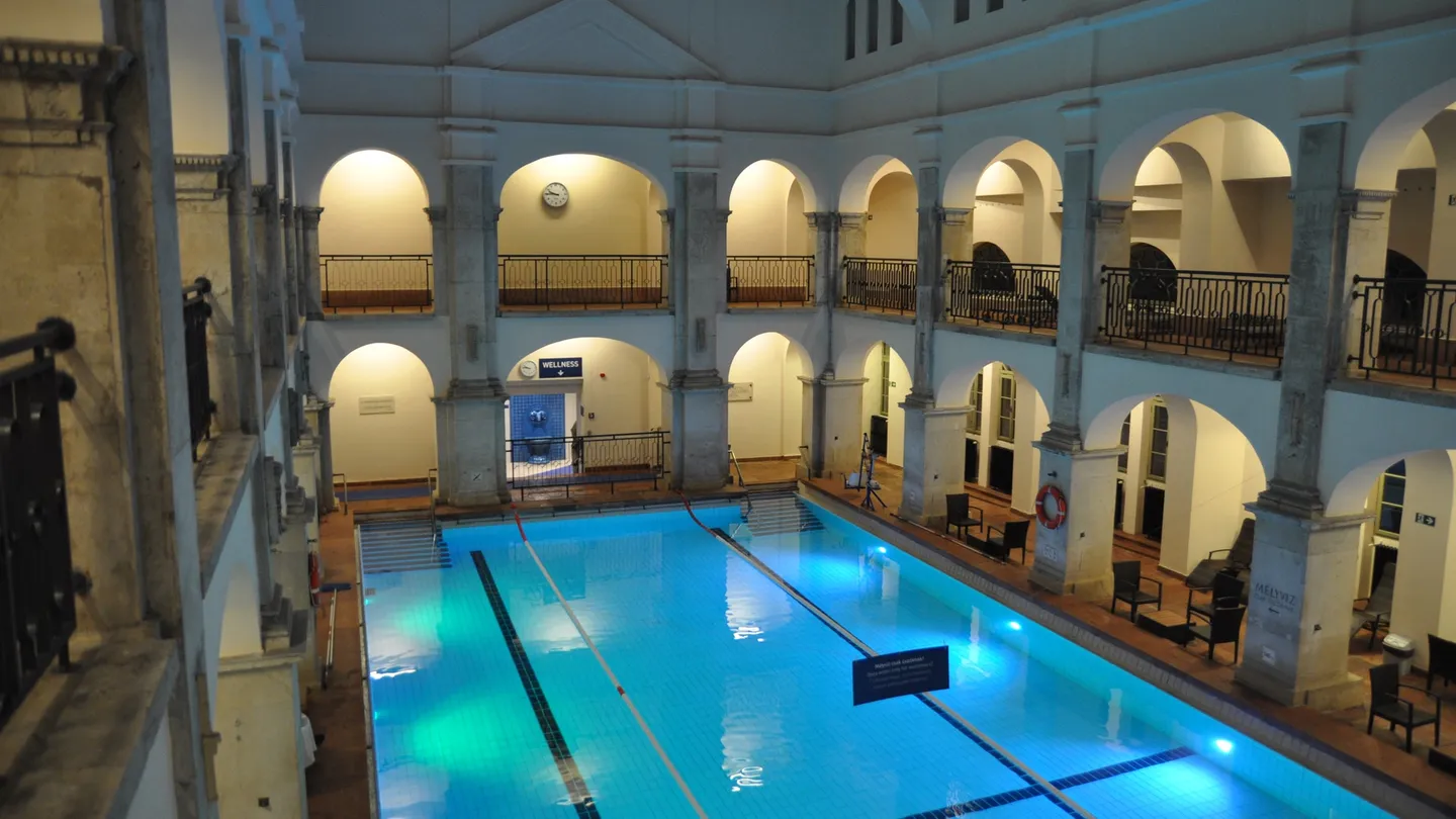 Piscina interna vazia dos Banhos Rudas, em Budapeste, com água azul iluminada e galerias de arcos em dois níveis