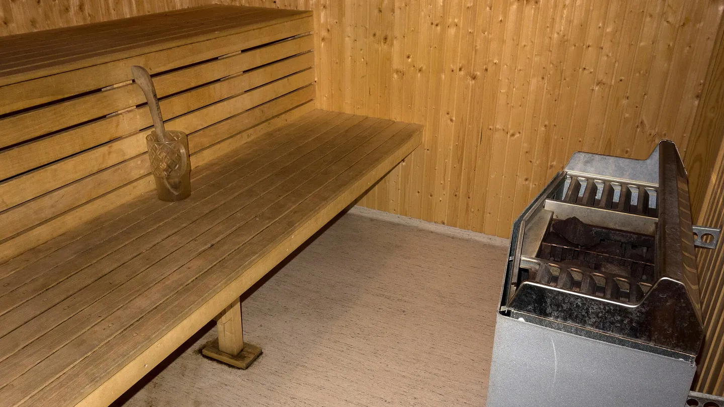 Interior de uma sauna revestida de madeira clara, com bancada em dois níveis à esquerda e forno elétrico com pedras à direita