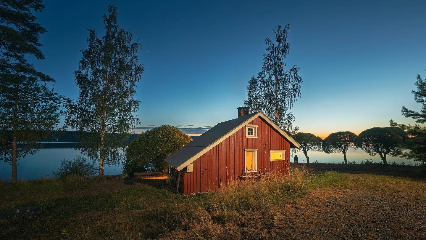 Cabana de sauna de madeira vermelha à beira do lago Ätäskö, em Kitee, na Finlândia, com luz acesa ao anoitecer