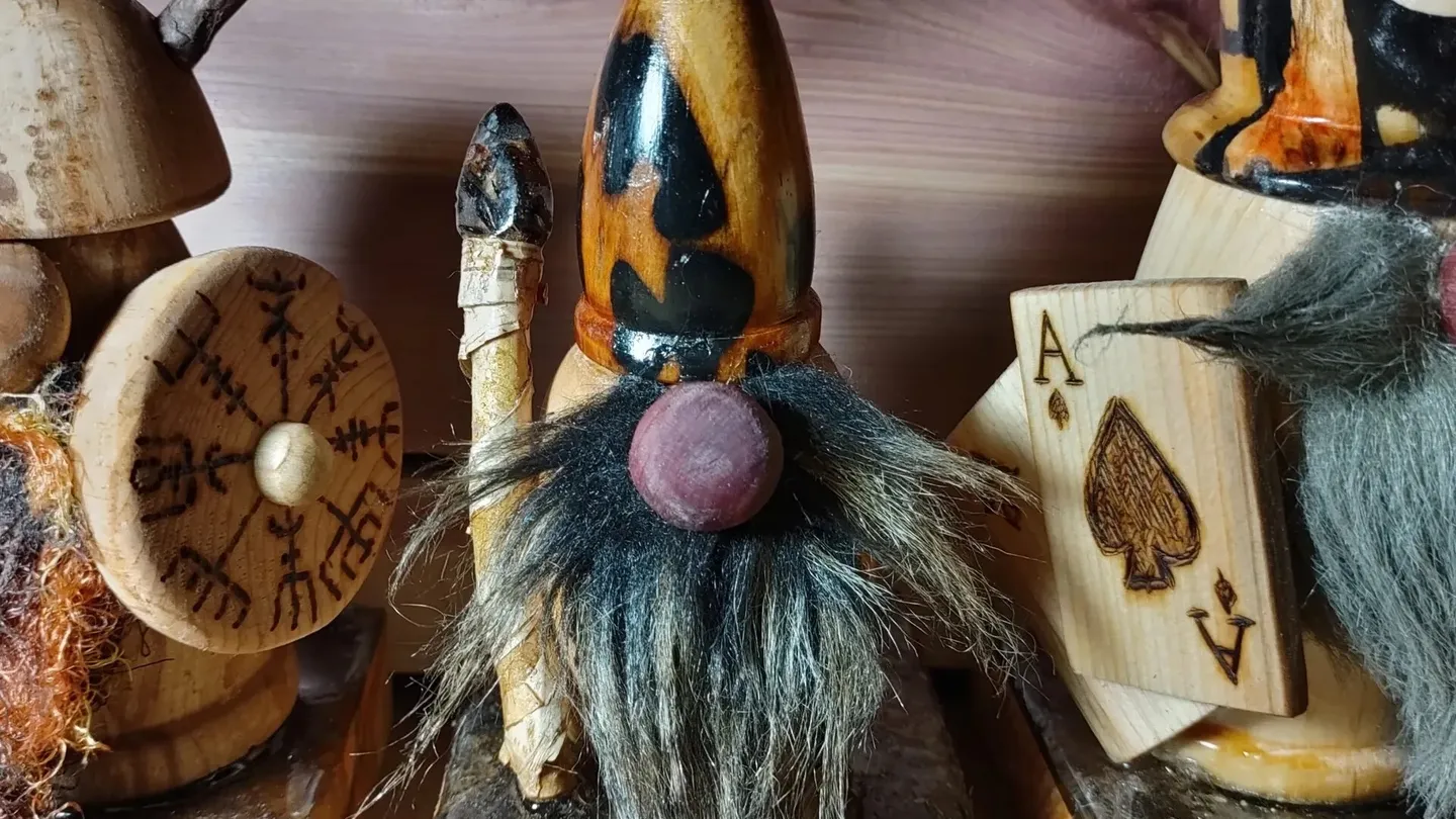 Três saunatonttu de madeira entalhada com barbas de pelo numa prateleira de madeira; o do meio, de gorro pontudo e nariz redondo, segura um cajado