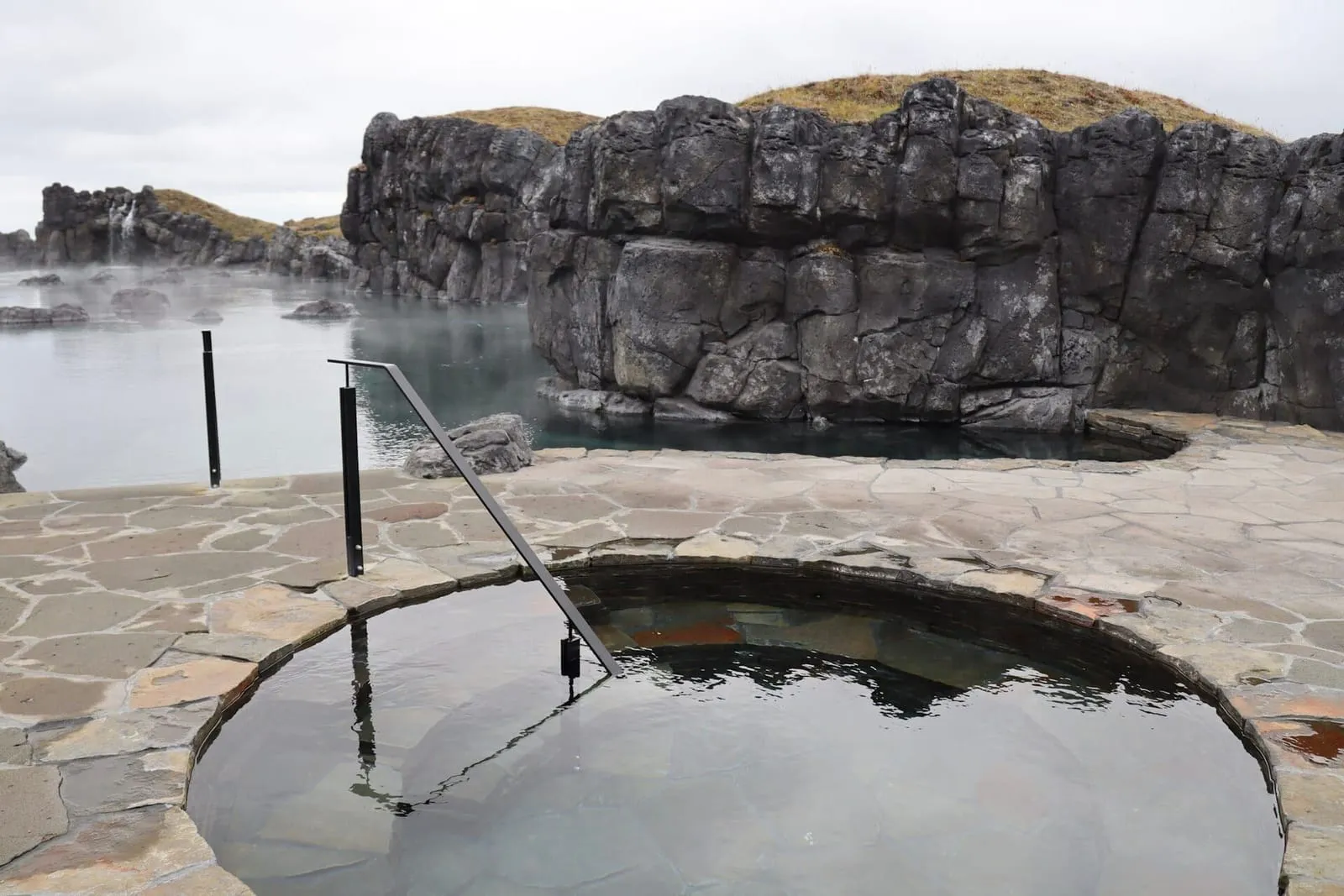 Piscina fria circular de pedra na Sky Lagoon, com muros de basalto e a lagoa geotermal ao fundo