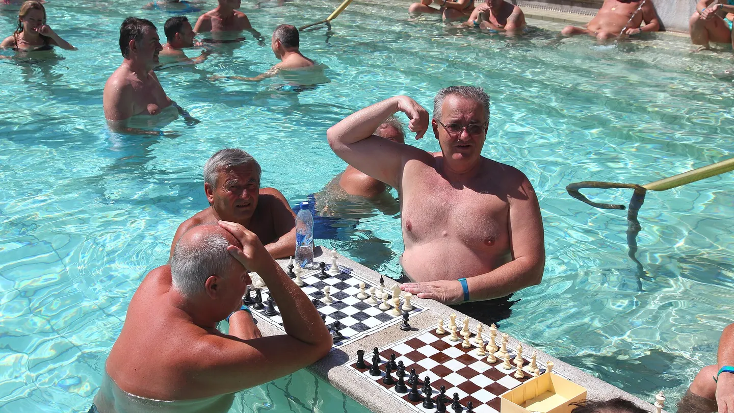 Homens jogando xadrez num tabuleiro apoiado na borda da piscina termal ao ar livre dos Banhos Széchenyi, em Budapeste, cercados por dezenas de banhistas